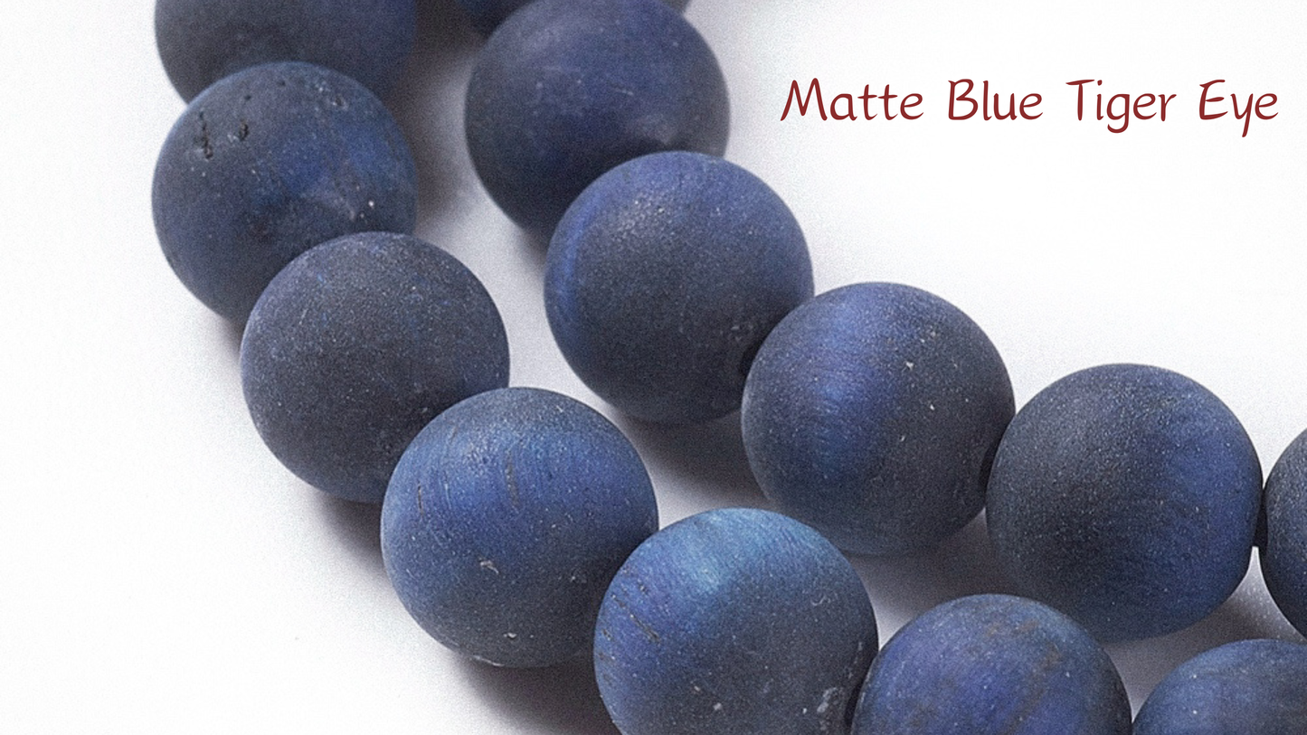 A macro close-up of Aking Sinta Matte Blue Tiger Eye beads showing a smooth, non-reflective velvet finish in deep navy and midnight blue; a white text label in the upper right corner reads "Matte Blue Tiger Eye."