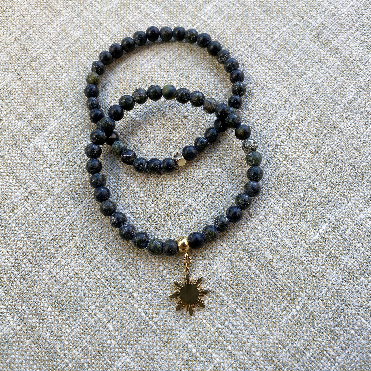 Aking Sinta Matte Serpentine Stretch Bracelet in gold, plain and with Araw charm, displayed on woven grey textured background, highlighting natural stone design