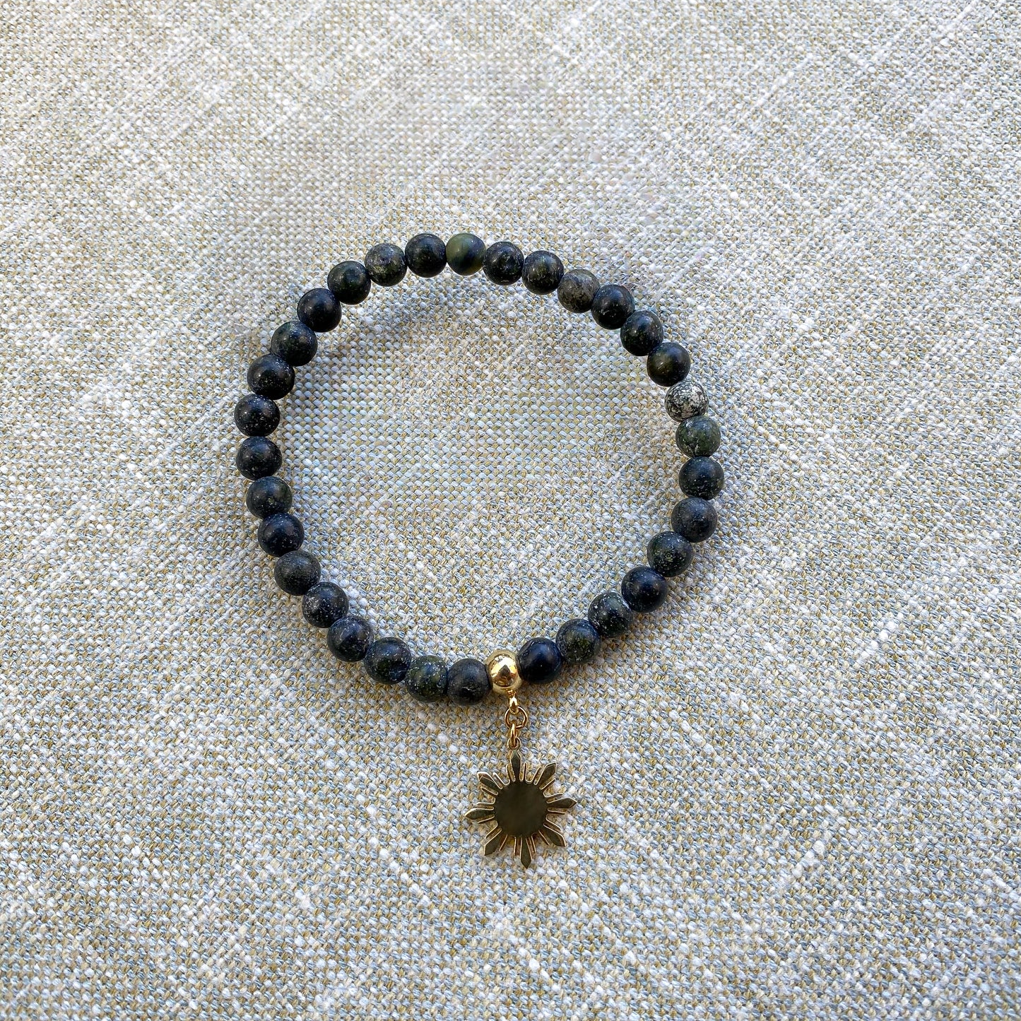 Aking Sinta Matte Serpentine Stretch Bracelet in gold with Araw charm displayed on woven grey textured background, showcasing natural stone beads and 8-ray sun motif