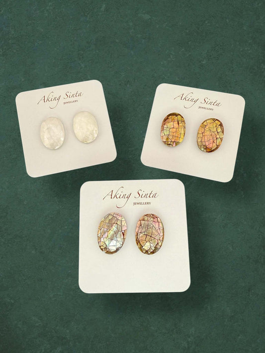 Three pairs of Aking Sinta oval mosaic mother of pearl stud earrings in white, gold, and iridescent pink hues. Handcrafted shell inlay jewelry presented on branded white display cards.