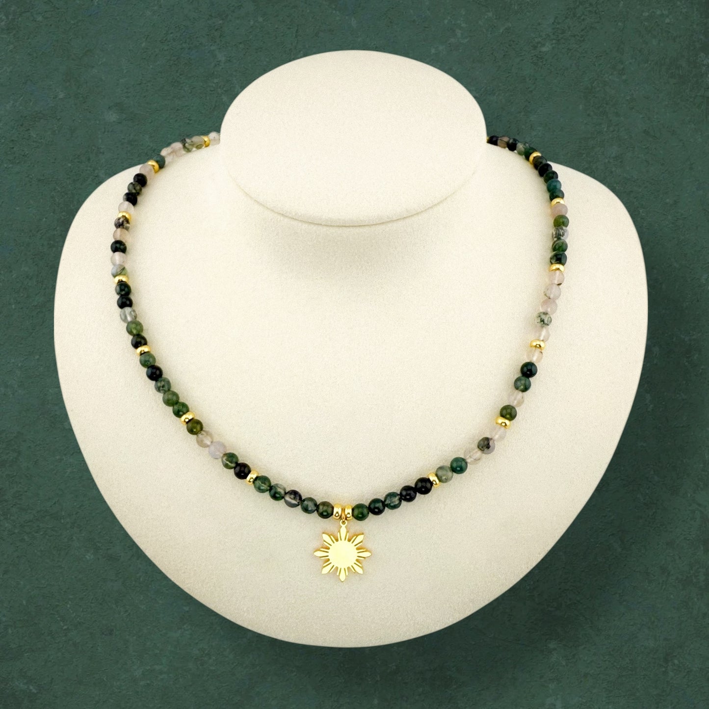 Aking Sinta Moss Agate beaded necklace on a white display bust; the strand features deep forest-green and translucent white gemstones with natural moss-like inclusions, accented by polished gold beads throughout the strand and a central 8-ray Araw sun charm.
