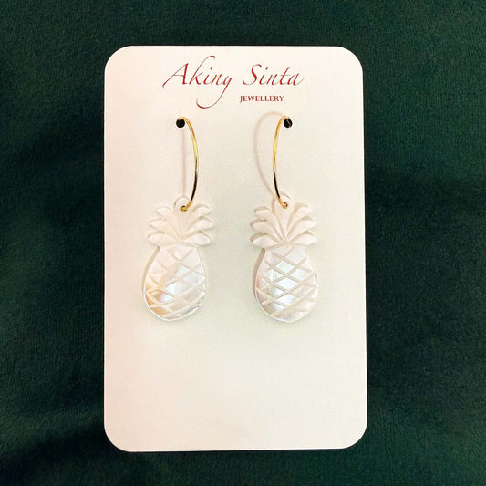 A pair of Aking Sinta pineapple earrings on a white branded display card. Each earring features a carved white mother of pearl pineapple charm hanging from a gold hoop, set against a dark green velvet background.