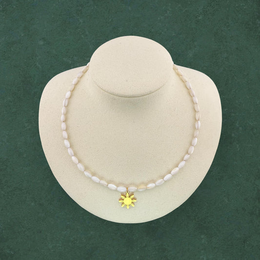 Aking Sinta Mother of Pearl rice bead necklace with a gold Araw (Sun) charm pendant, displayed on a black jewelry bust against a dark green background.
