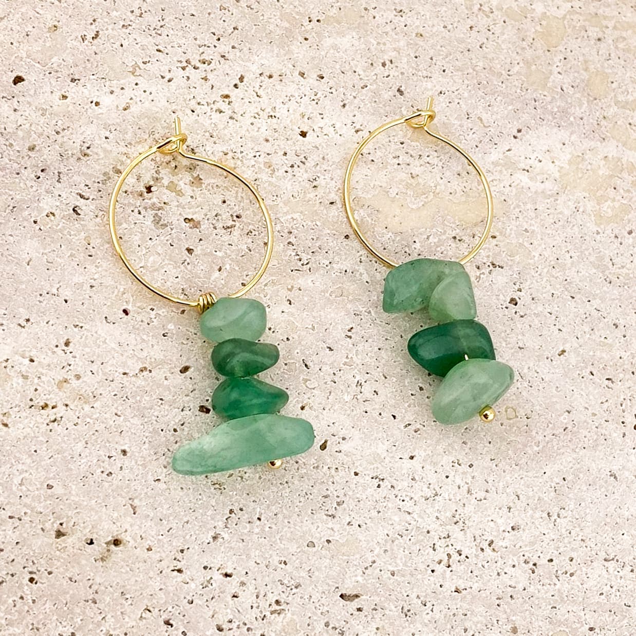 A pair of gold drop earrings featuring clusters of multiple small, raw green aventurine nuggets, resting on a gray marble slab; the unpolished texture of the stones contrasts with the smooth, veined marble surface.