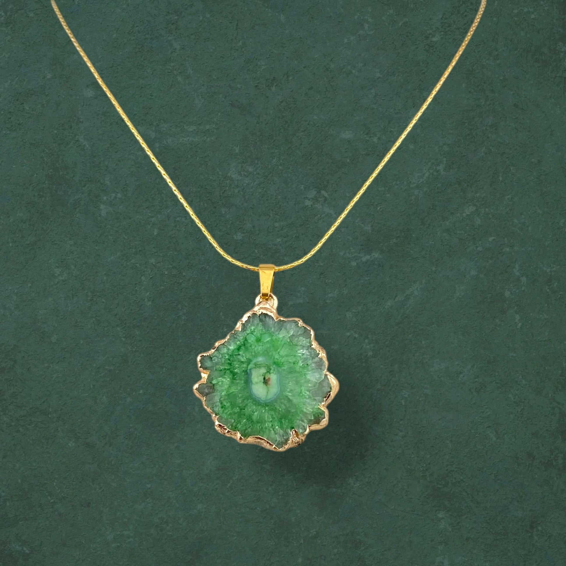 A natural Aking Sinta Druzy Agate pendant necklace on a solid dark green background; the organic-shaped stone features a sparkling crystalline center and is suspended from a delicate gold chain.