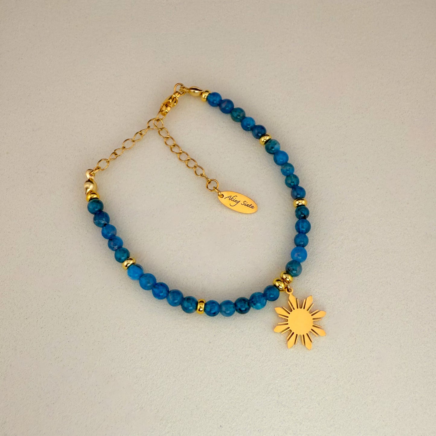Aking Sinta Apatite beaded bracelet featuring vibrant neon-blue gemstones, a gold 8-ray Araw sun charm, and a gold extender chain with a teardrop-shaped tag engraved with "Aking Sinta" branding; displayed on a crisp white background.