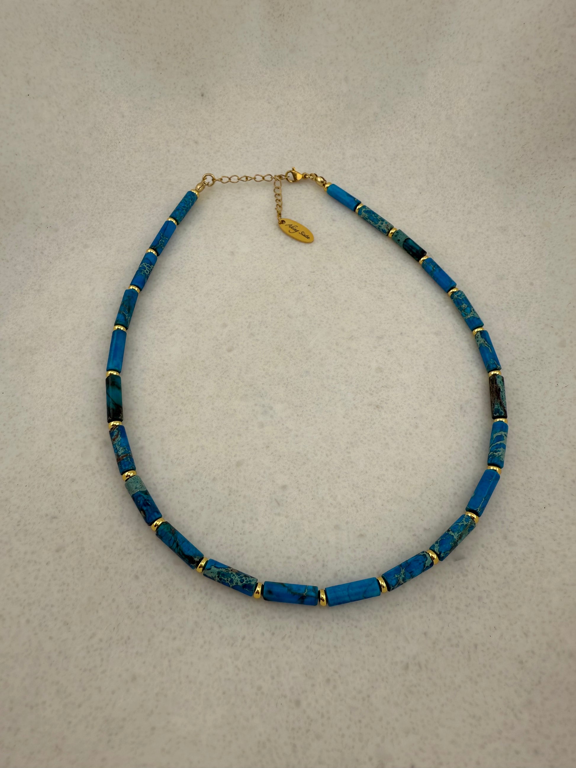 Aking Sinta Ocean Blue Imperial Jasper necklace with long column beads and gold spacers on a white background; includes a gold teardrop pendant engraved with "Aking Sinta" at the clasp.