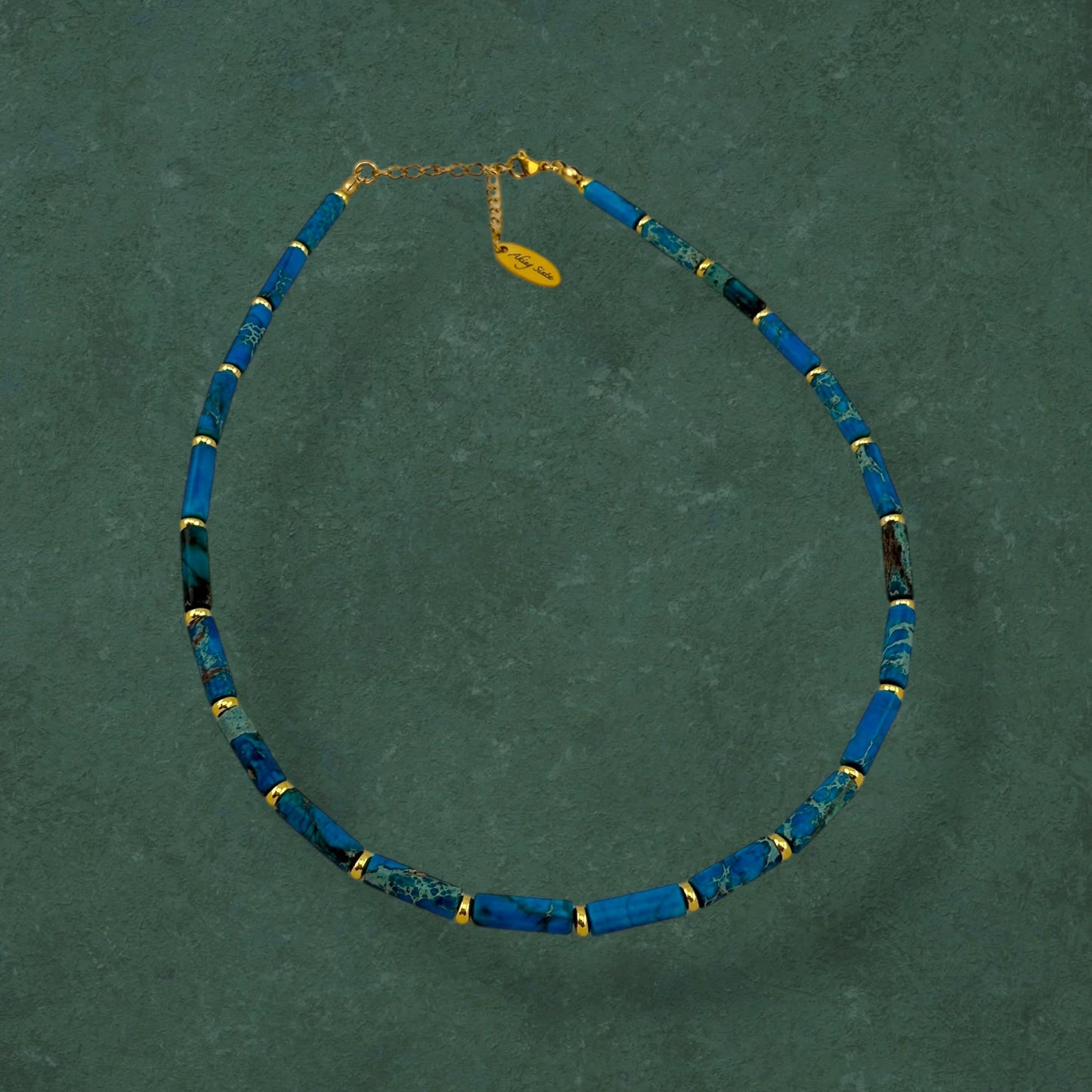 Aking Sinta Ocean Blue Imperial Jasper necklace with long column beads and gold spacers on a dark green background; featuring a gold teardrop-shaped pendant engraved with "Aking Sinta" at the clasp.
