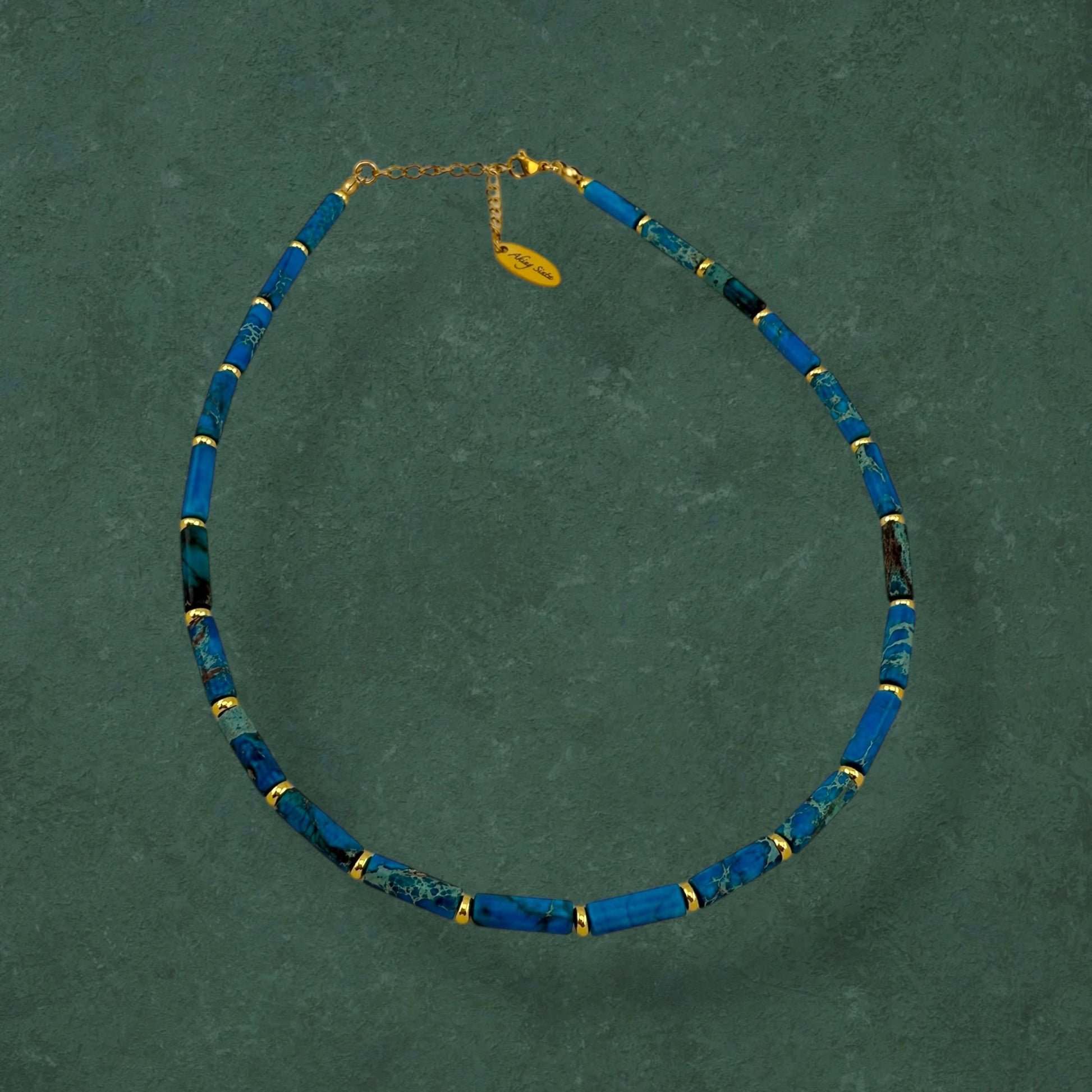 Aking Sinta Ocean Blue Imperial Jasper necklace with long column beads and gold spacers on a dark green background; featuring a gold teardrop-shaped pendant engraved with "Aking Sinta" at the clasp.