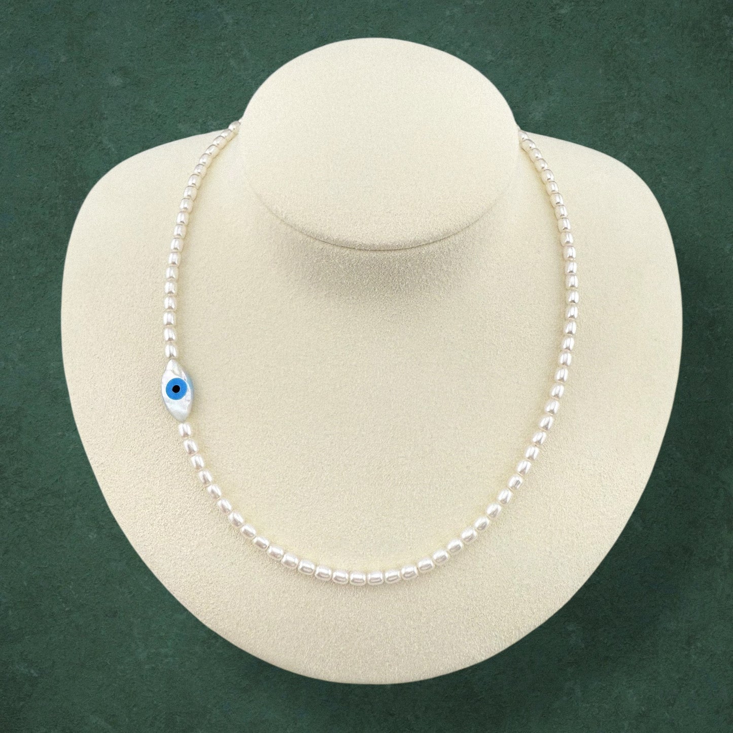 Aking Sinta gold necklace with an asymmetrical off-centre Mother of Pearl Evil Eye pendant, displayed on a white jewelry bust against a dark green background.