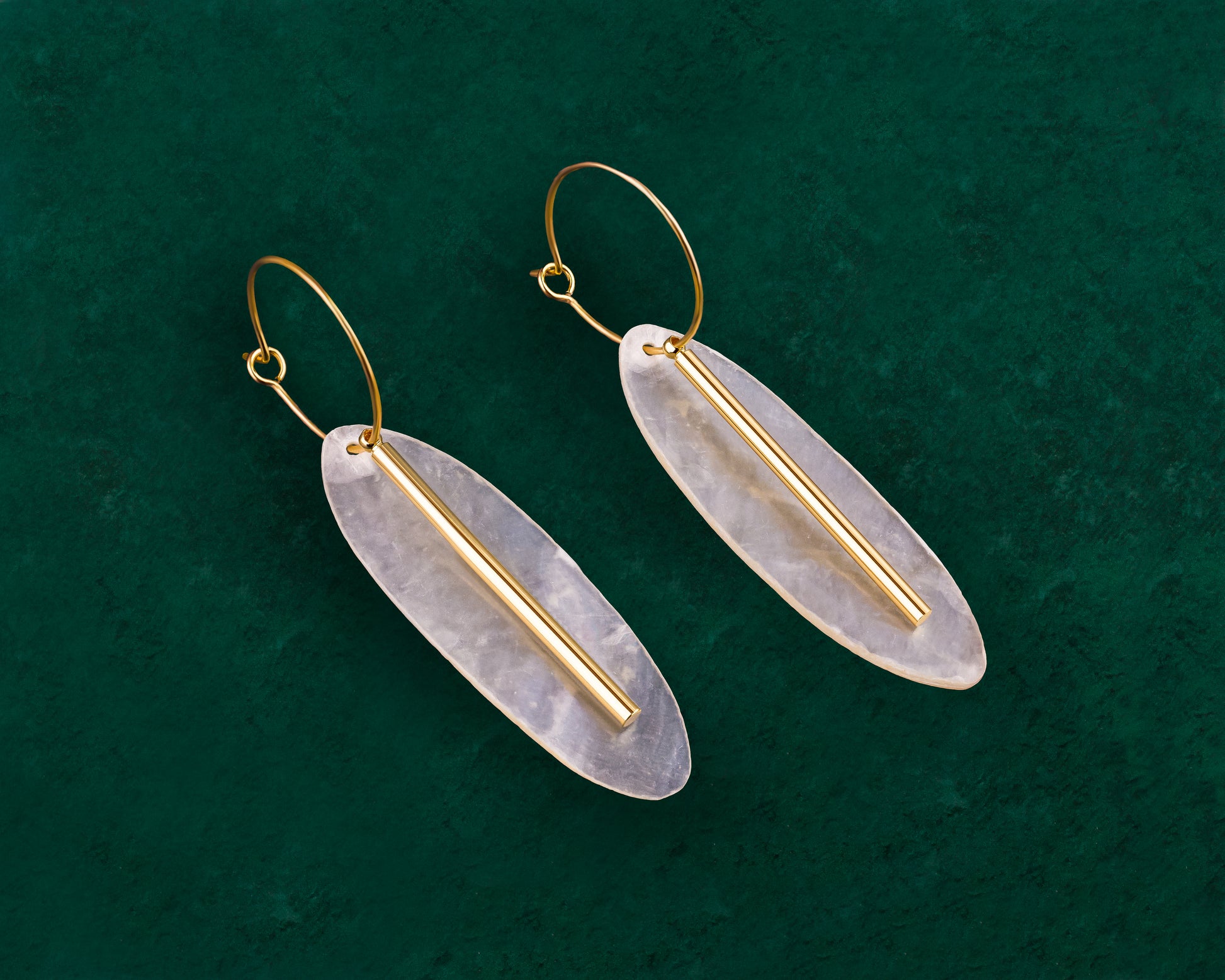 A pair of Aking Sinta gold hoop earrings on a dark green background; each features a vertically oriented, oval-shaped white Capiz shell disk accented with a slim, polished gold metal bar running down the center.