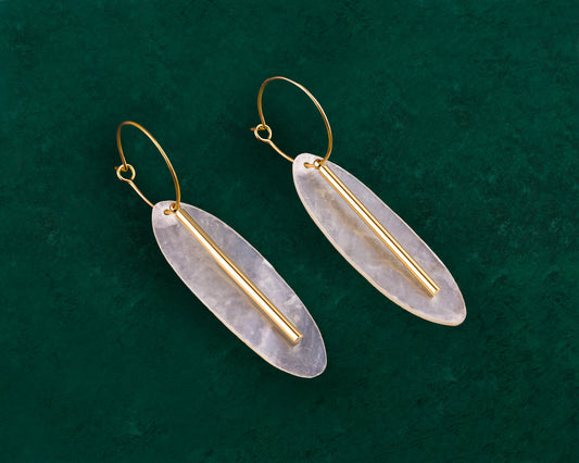 A pair of Aking Sinta gold hoop earrings on a dark green background; each features a vertically oriented, oval-shaped white Capiz shell disk accented with a slim, polished gold metal bar running down the center.