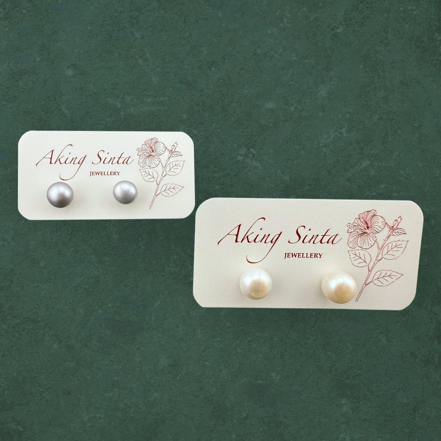 A side-by-side display of Aking Sinta Palawan Pearl Studs featuring one pair of classic white pearls and one pair of peacock-black pearls on a branded jewelry card.