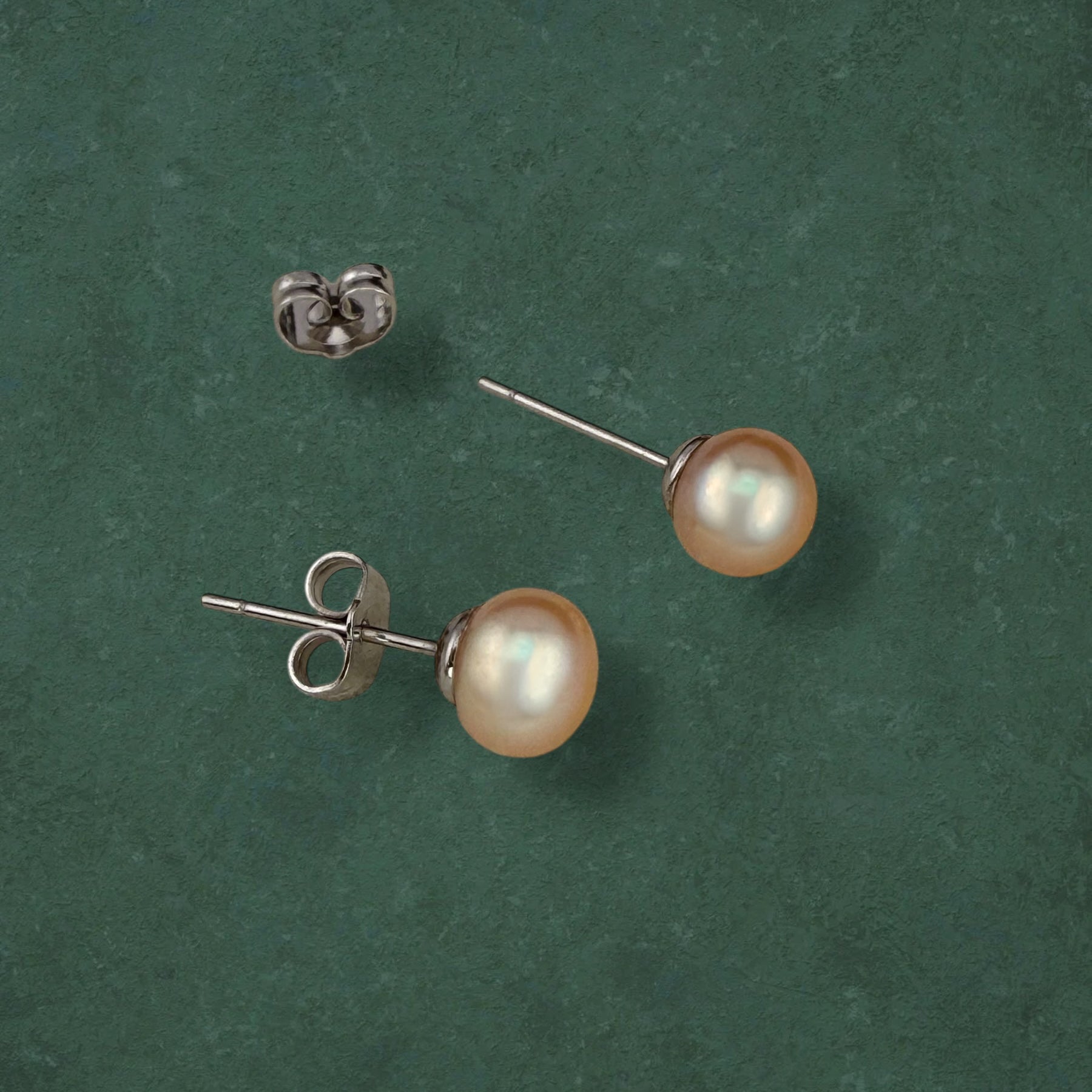 Close-up of an Aking Sinta Peach Palawan Pearl stud showing the gold butterfly lock and natural peach luster against a dark green background.