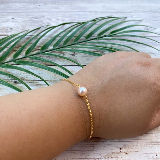 A minimalist Aking Sinta bracelet featuring a single peach-colored freshwater pearl on a delicate gold chain. Shown being worn on a wrist against a beach-style wood background with a green palm leaf