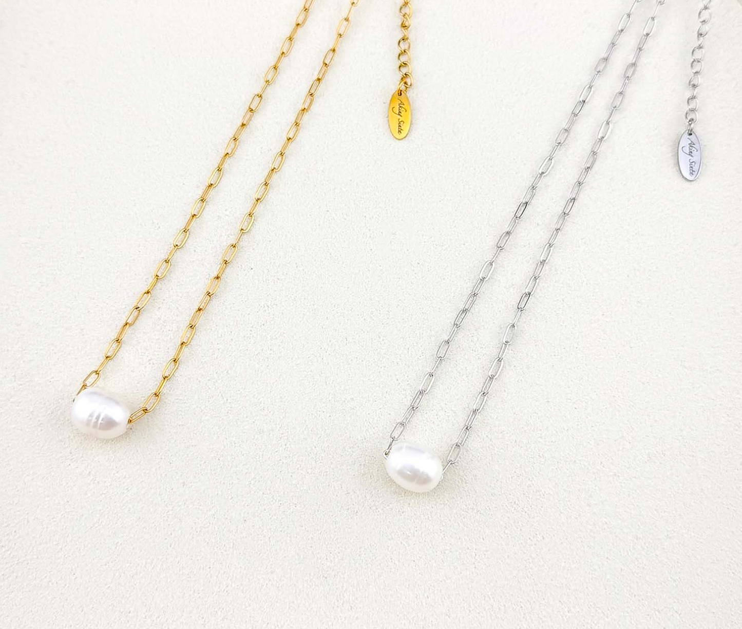 Minimalist Aking Sinta necklaces featuring floating white baroque pearls on gold and silver paperclip chains. Close-up of elegant link jewelry on a white textured surface.