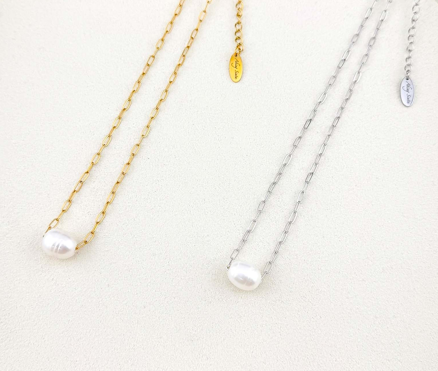 Minimalist Aking Sinta necklaces featuring floating white baroque pearls on gold and silver paperclip chains. Close-up of elegant link jewelry on a white textured surface.