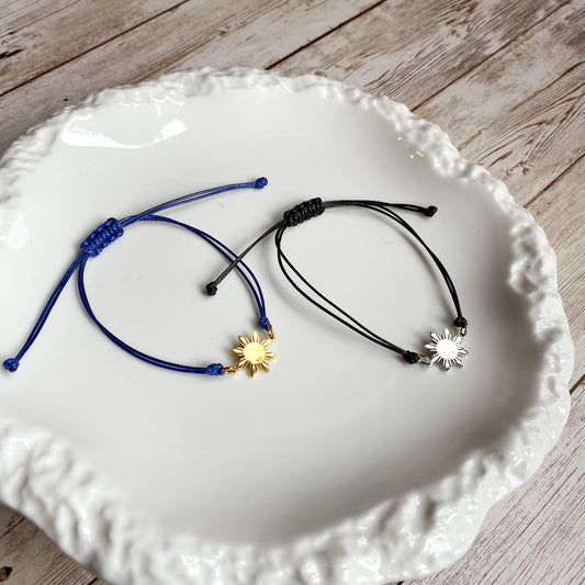 A pair of Aking Sinta Simple Philippine Sun Unisex Friendship Bracelets featuring a silver 8-ray Araw charm and a gold 8-ray Araw charm; both displayed inside a shimmering, iridescent Mother of Pearl shell container for a luxury coastal aesthetic.