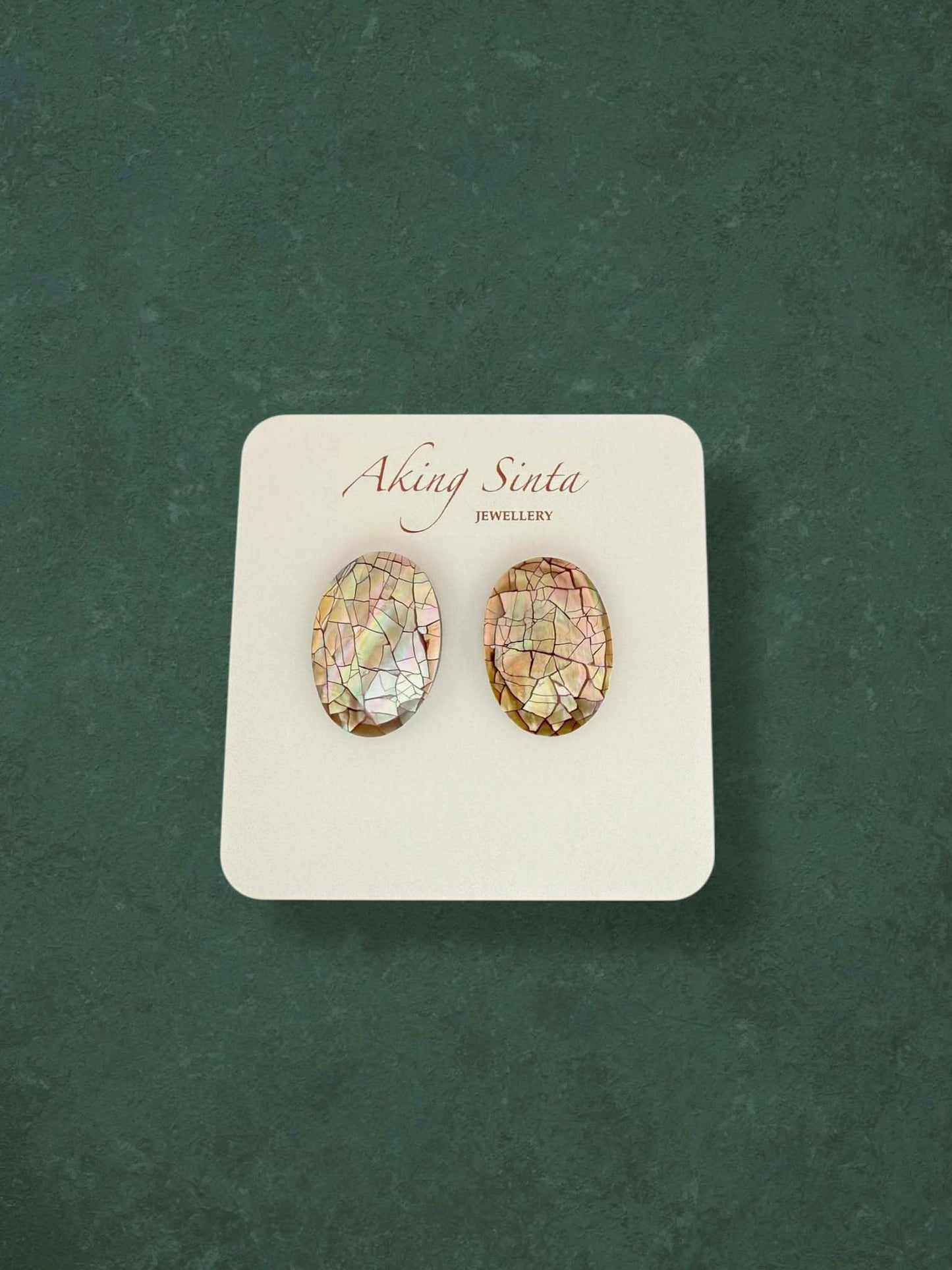 A pair of Aking Sinta oval mosaic mother of pearl stud earrings featuring iridescent pink, green, and brown tones. These handcrafted shell inlay earrings showcase a shimmering cracked mosaic effect on a branded white card