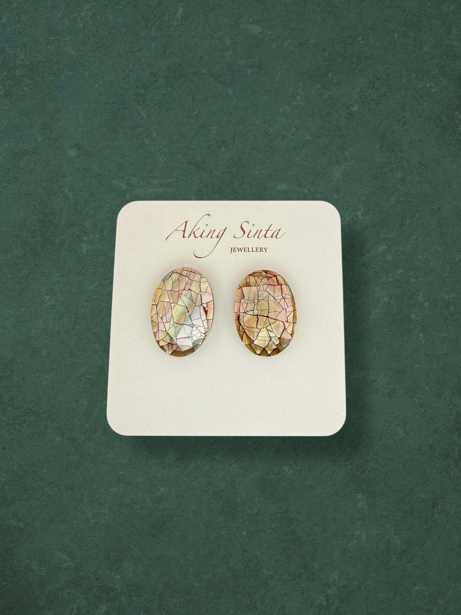 A pair of Aking Sinta oval mosaic mother of pearl stud earrings featuring iridescent pink, green, and brown tones. These handcrafted shell inlay earrings showcase a shimmering cracked mosaic effect on a branded white card