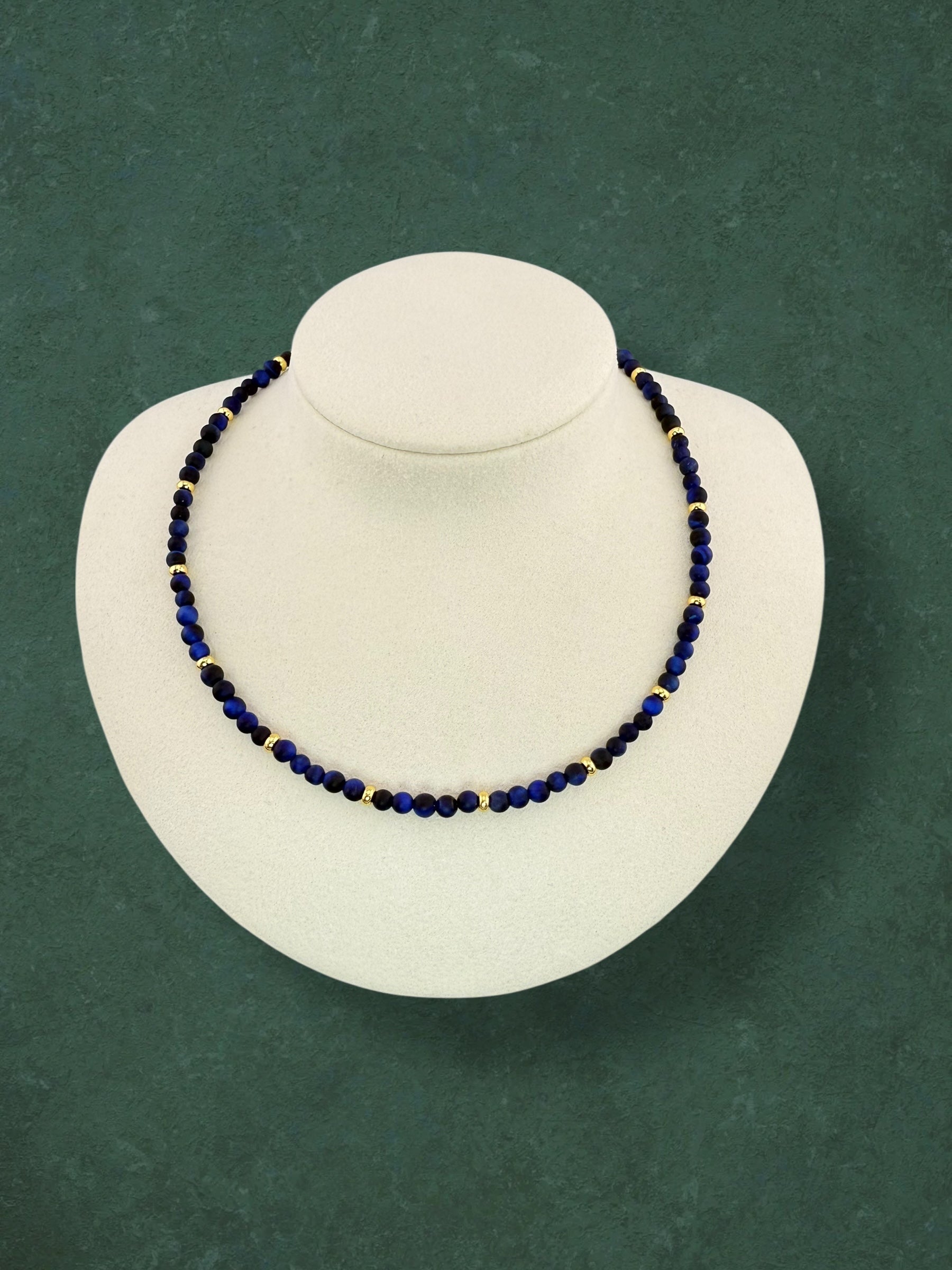 A plain Aking Sinta Blue Tiger Eye beaded necklace without a charm, displayed on a dark green background; the dark blue stones feature a natural shimmering stripe known as chatoyancy.