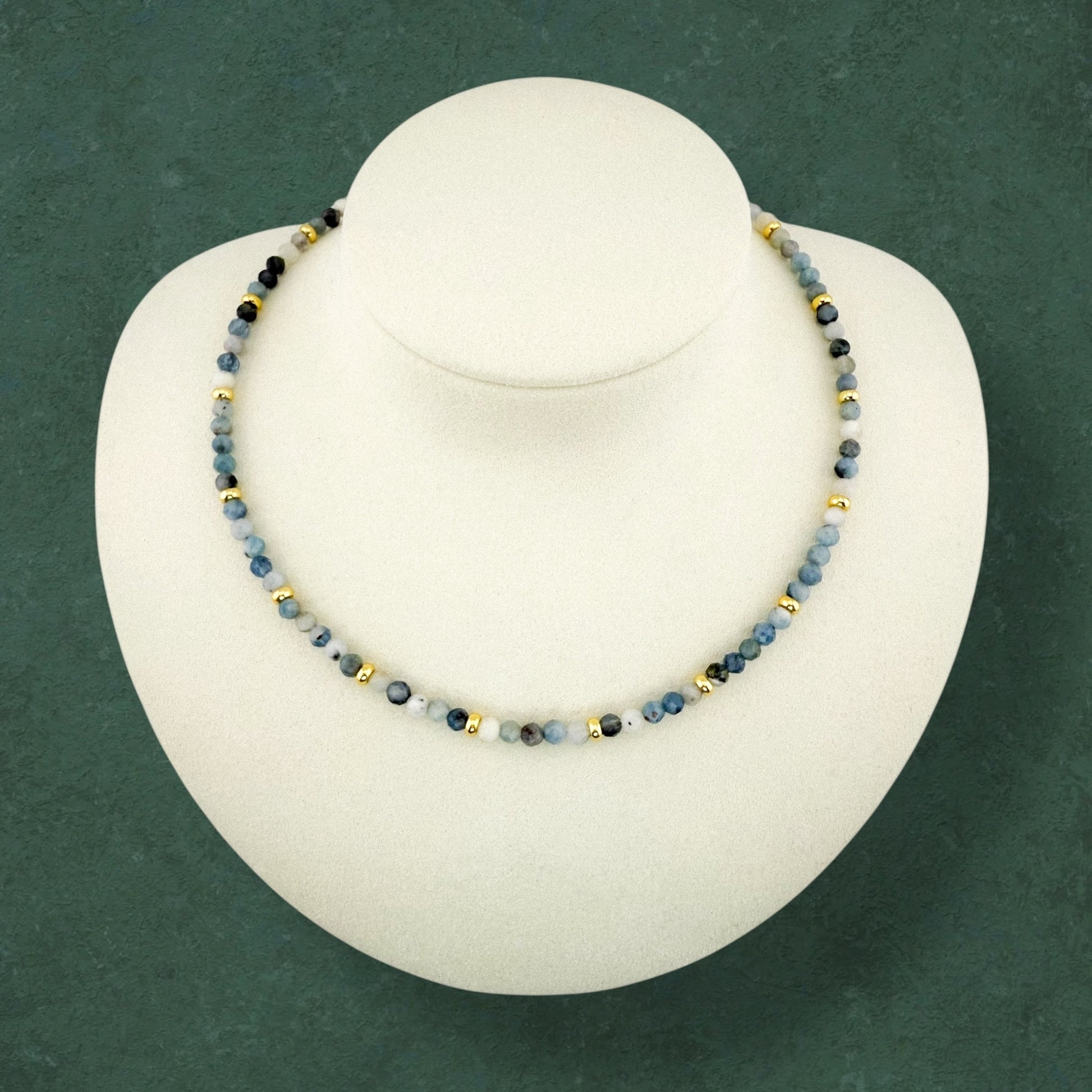A plain Aking Sinta Dirty Aquamarine beaded necklace on a white display bust; featuring a mix of light and dark blue beads punctuated with small gold accent beads without a central pendant.