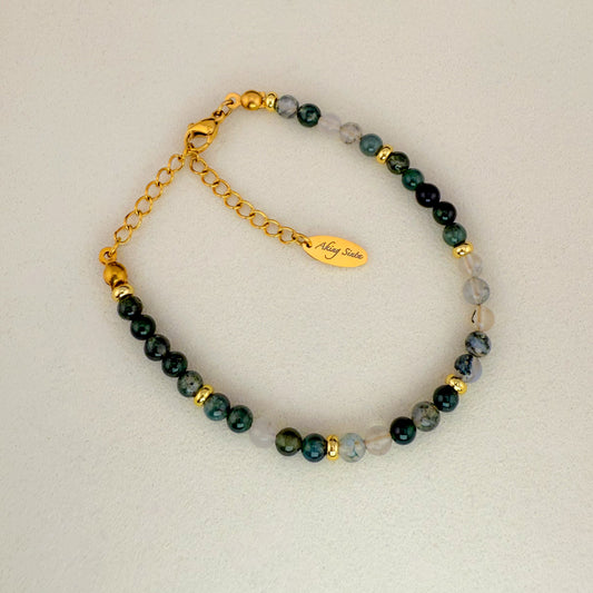 A minimalist Aking Sinta Moss Agate beaded bracelet without a charm, laid on a white background; featuring forest-green stones and a gold extender chain with a teardrop-shaped metal tag engraved with the "Aking Sinta" brand name.