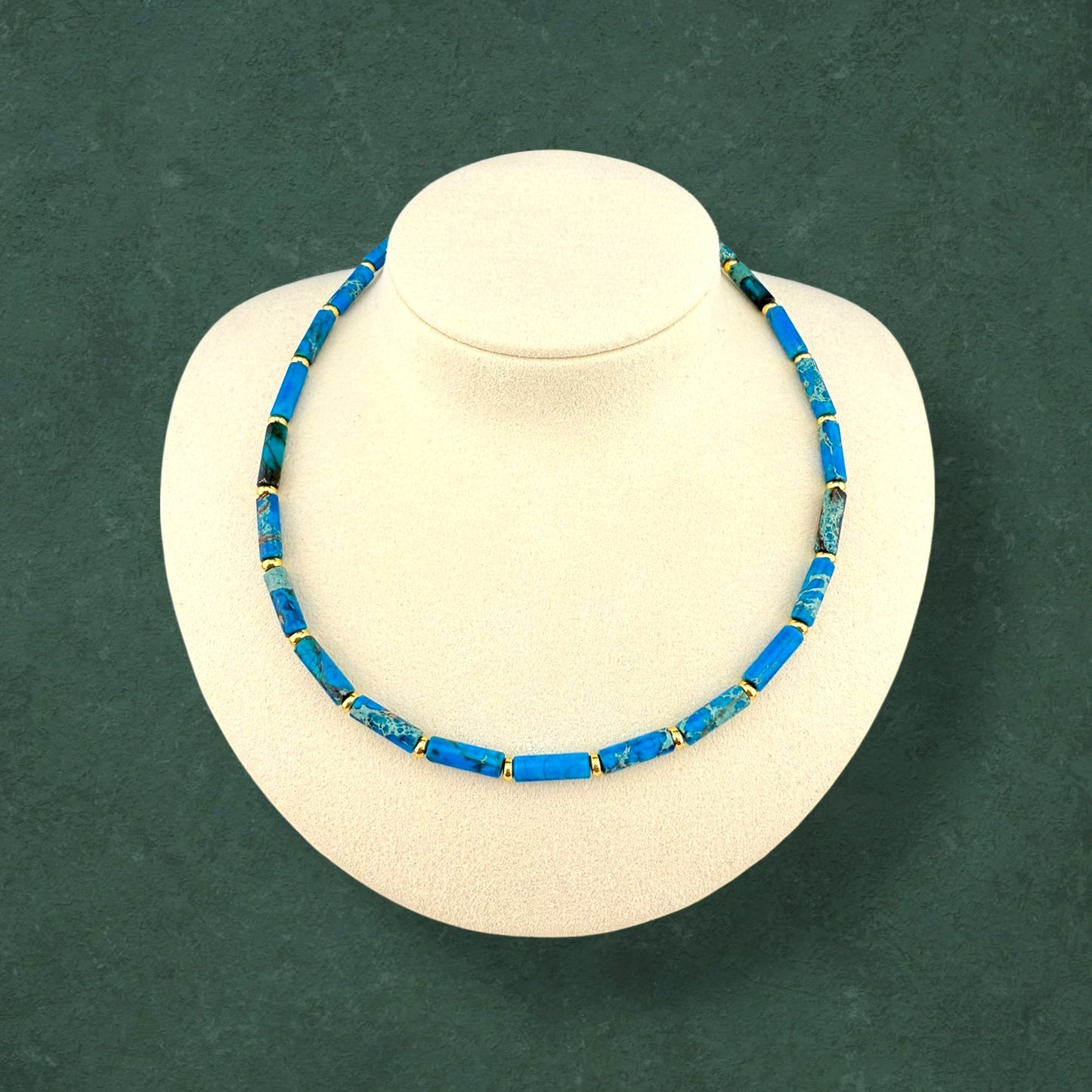 A plain Aking Sinta Ocean Blue Imperial Jasper necklace without a charm; featuring long column-shaped blue beads with a small gold accent bead between each one, displayed on a white bust.