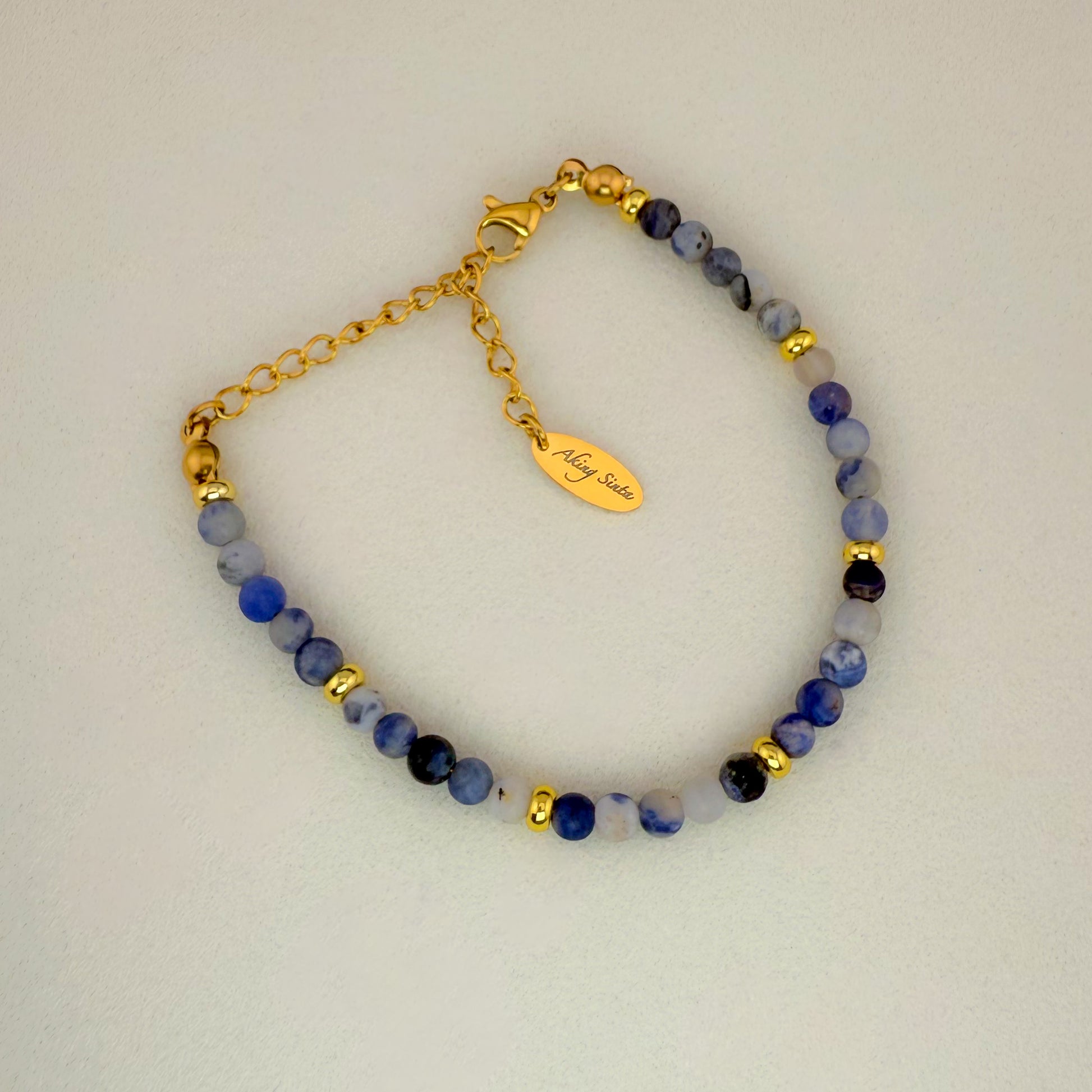 A minimalist Aking Sinta Sodalite beaded bracelet without a charm, featuring navy blue gemstone beads with natural white veins and a gold extender chain with a teardrop-shaped metal tag engraved with the "Aking Sinta" brand name.