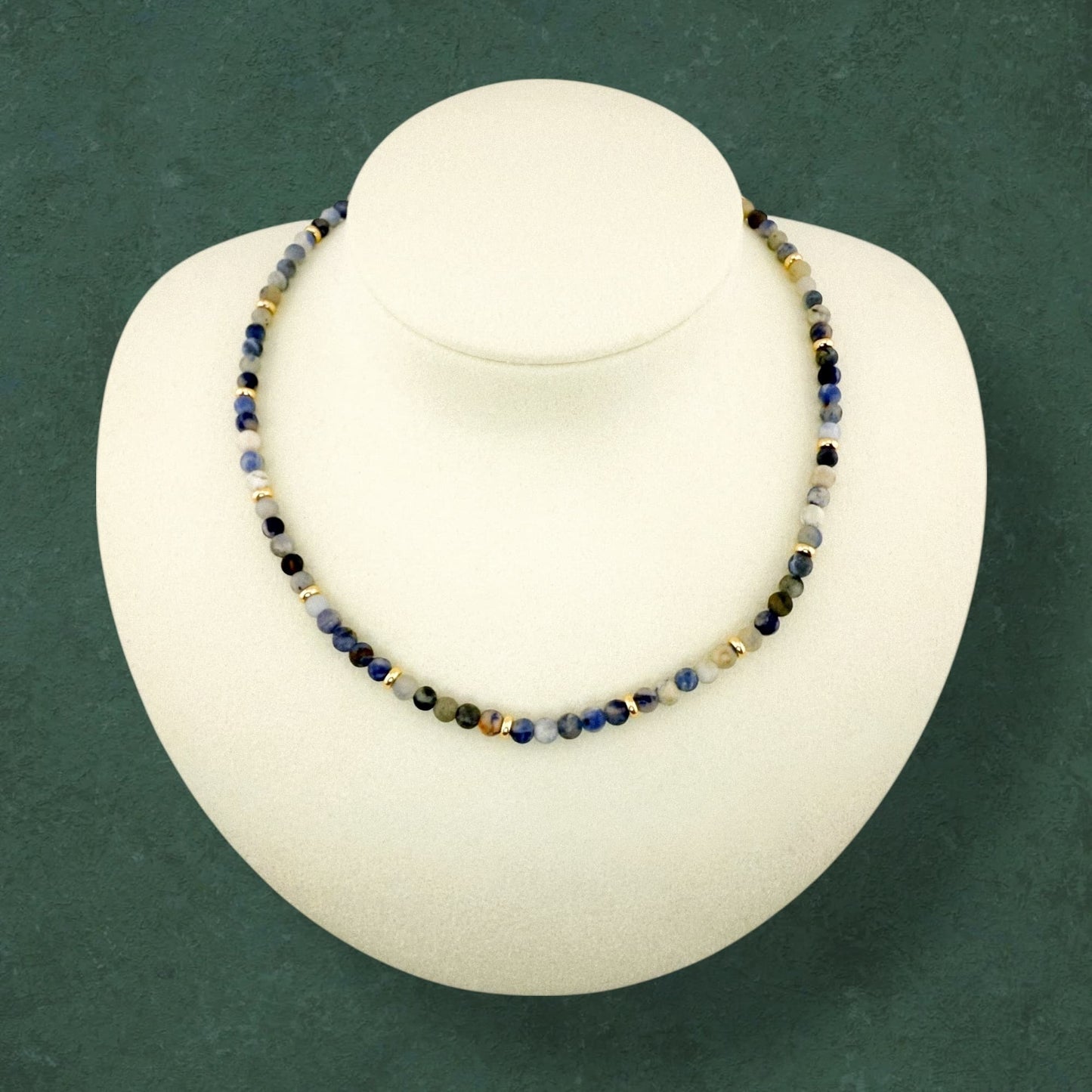 A plain Aking Sinta Sodalite necklace without a charm, draped on a white jewelry bust; featuring white, cream, and light green beads with small gold accent beads.