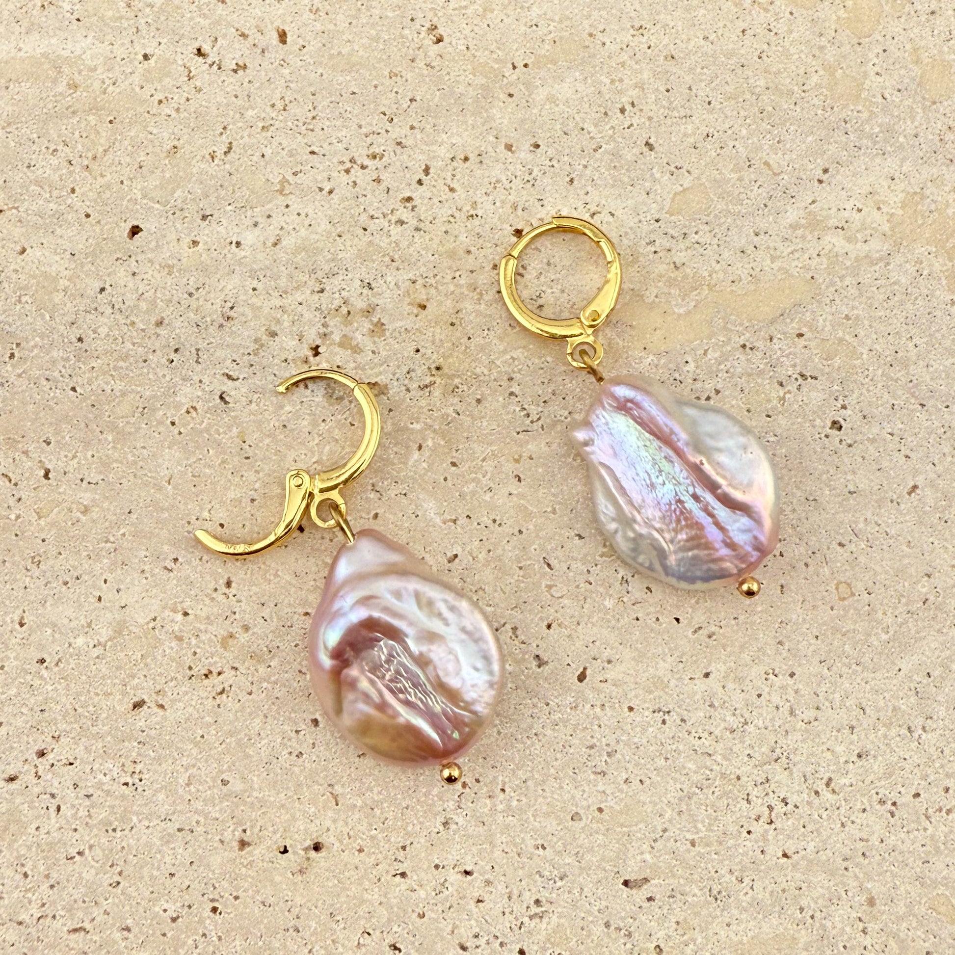 Close-up of Aking Sinta purple Keshi pearl huggie earrings on a white stone; one gold hoop is open to show the hinge clasp mechanism.