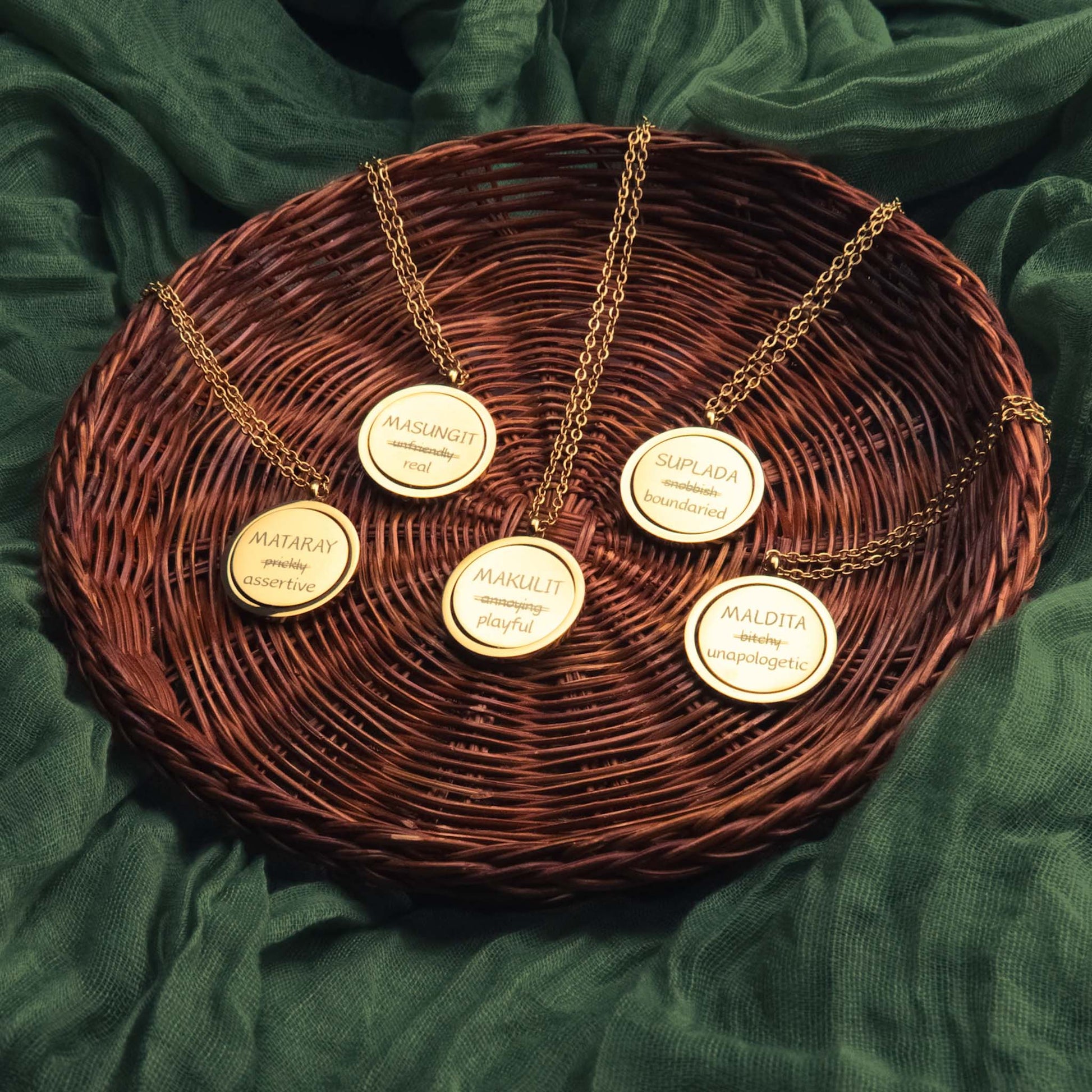 A collection of five gold Aking Sinta spinning coin fidget necklaces arranged in a woven basket. Each pendant features a reclaimed Tagalog word with its traditional negative meaning crossed out and replaced by an empowering one: Mataray (assertive), Masungit (real), Makulit (playful), Suplada (boundaried), and Maldita (unapologetic). The necklaces are set against a dark green fabric backdrop.