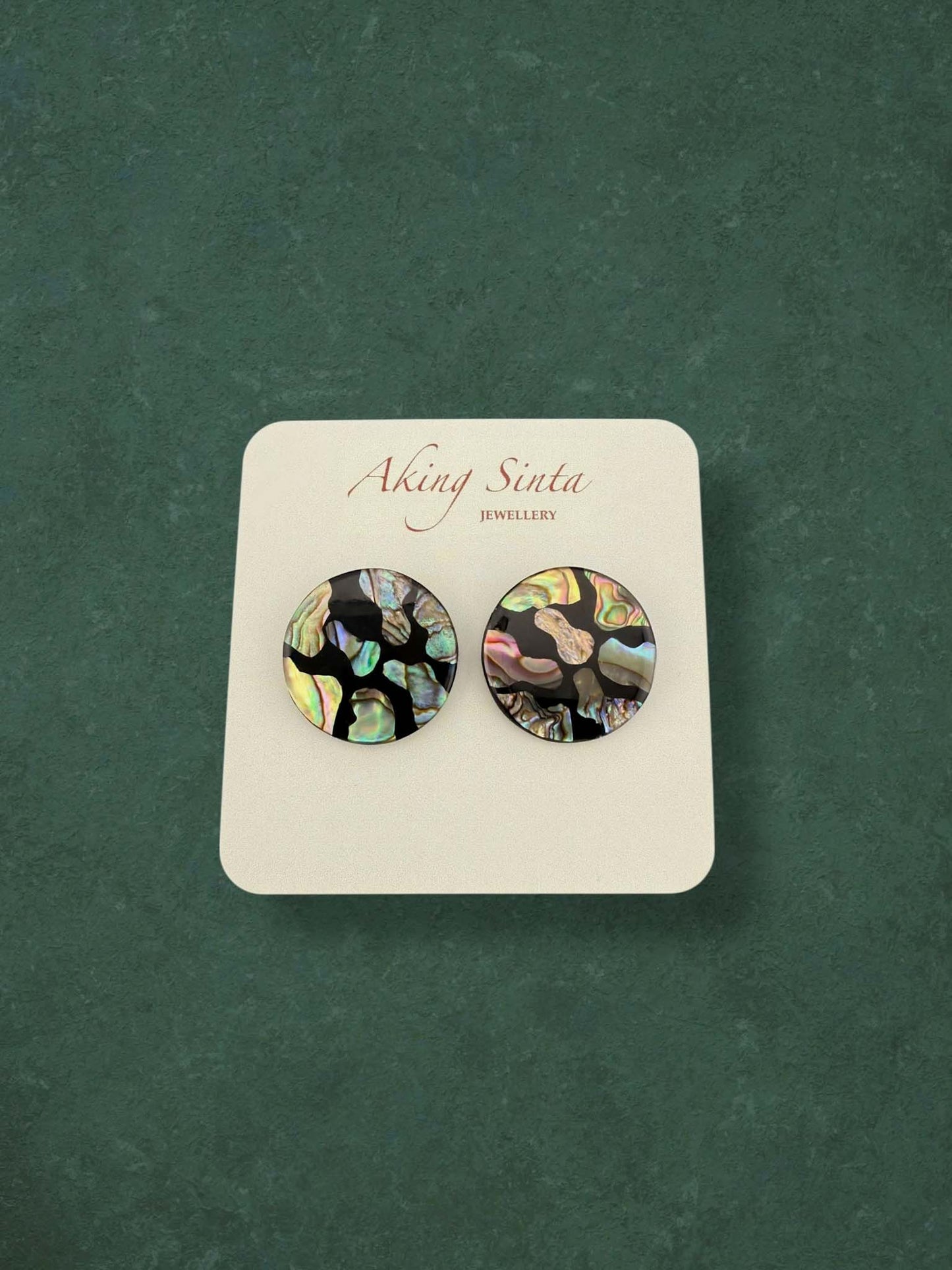 A pair of Aking Sinta round abalone shell stud earrings featuring a dark mosaic design. The iridescent shell fragments display vibrant peacock-green and pink hues against a black base, presented on a branded card