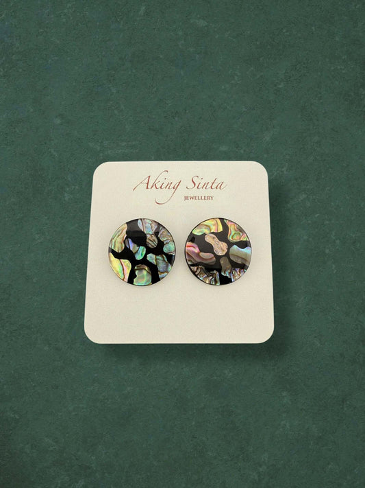 A pair of Aking Sinta round abalone shell stud earrings featuring a dark mosaic design. The iridescent shell fragments display vibrant peacock-green and pink hues against a black base, presented on a branded card