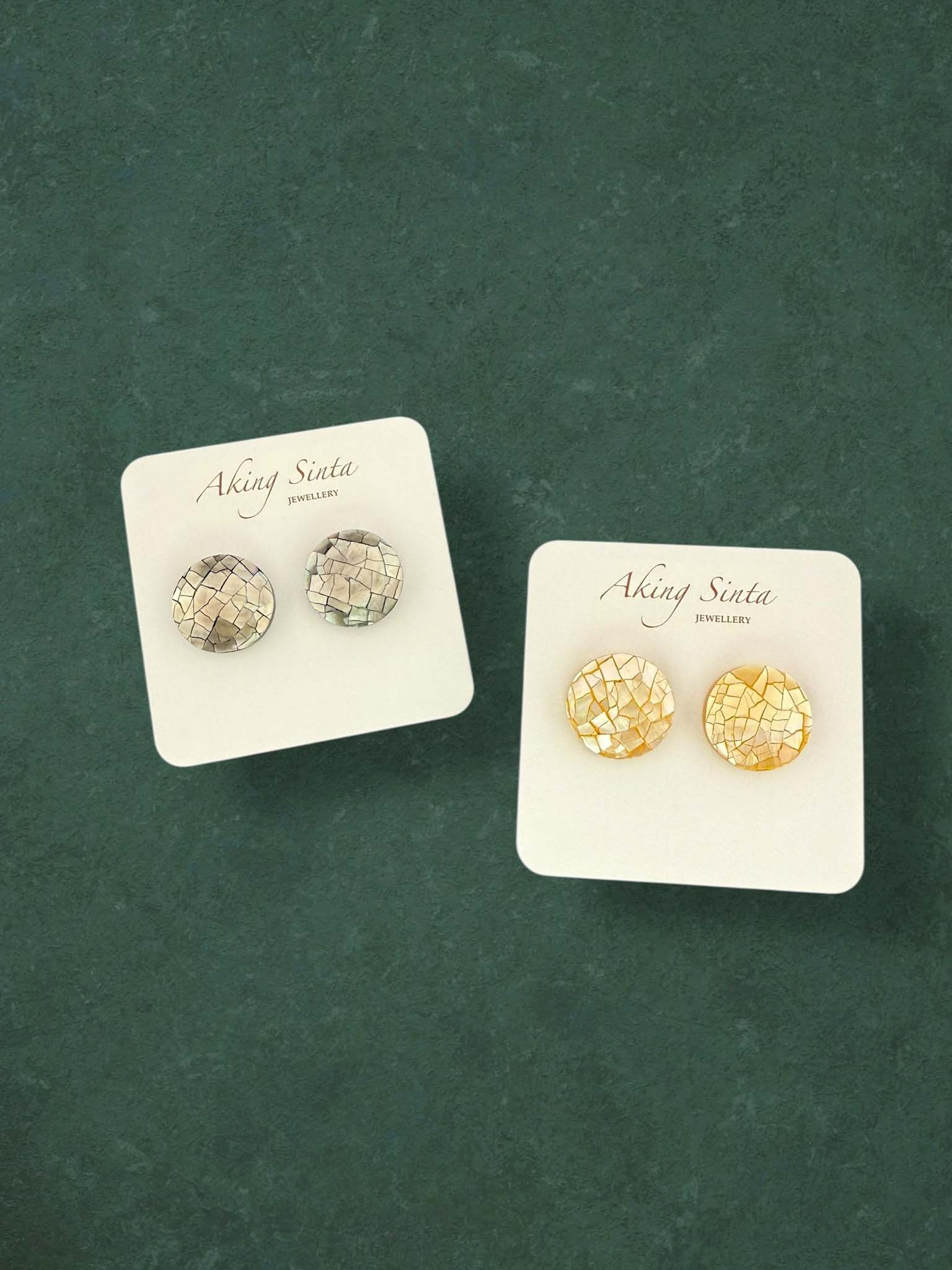 Two pairs of Aking Sinta round mosaic mother of pearl stud earrings in iridescent grey and golden yellow. These circular shell inlay earrings are displayed on branded cards against a dark green background