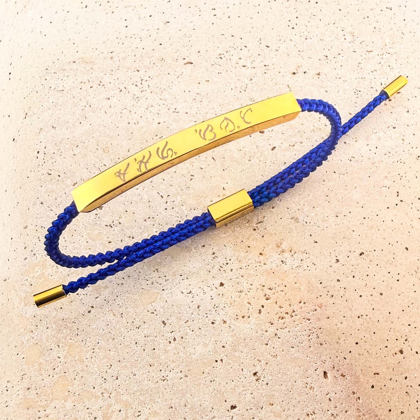 An Aking Sinta friendship bracelet featuring a royal blue woven cord and a polished gold bar. The bar is custom-engraved with ancient Filipino Baybayin script. The adjustable bracelet is displayed on a light-colored natural stone surface with gold-toned end caps.