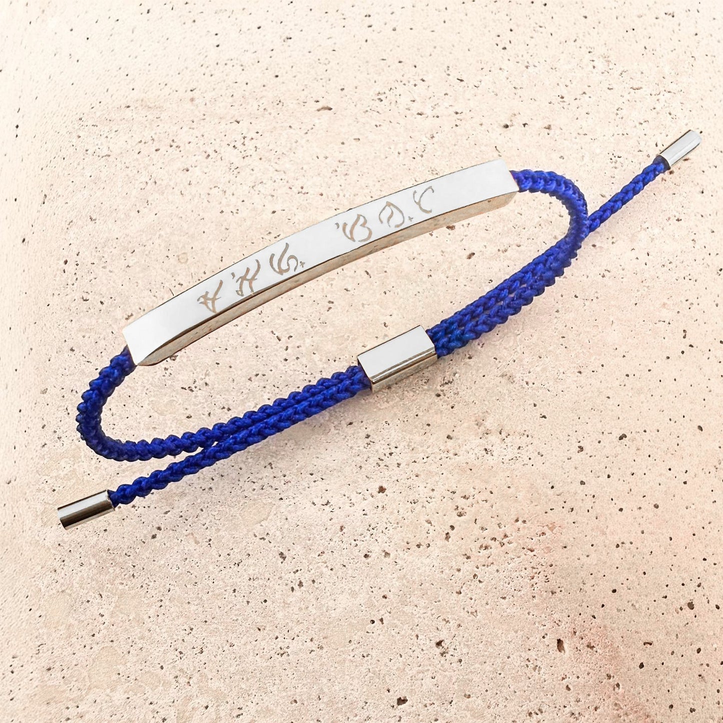 An Aking Sinta friendship bracelet featuring a royal blue woven cord and a sleek silver bar. The bar is custom-engraved with ancient Filipino Baybayin script. This adjustable cord bracelet is finished with silver-toned end caps and shown on a light-colored natural stone background.
