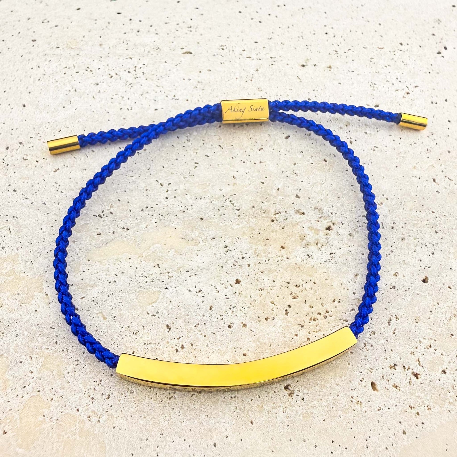 An Aking Sinta friendship bracelet featuring a vibrant royal blue woven cord and a curved, polished gold bar. The adjustable bracelet includes a gold-toned slider engraved with the "Aking Sinta" logo and matching end caps, displayed on a light natural stone surface.