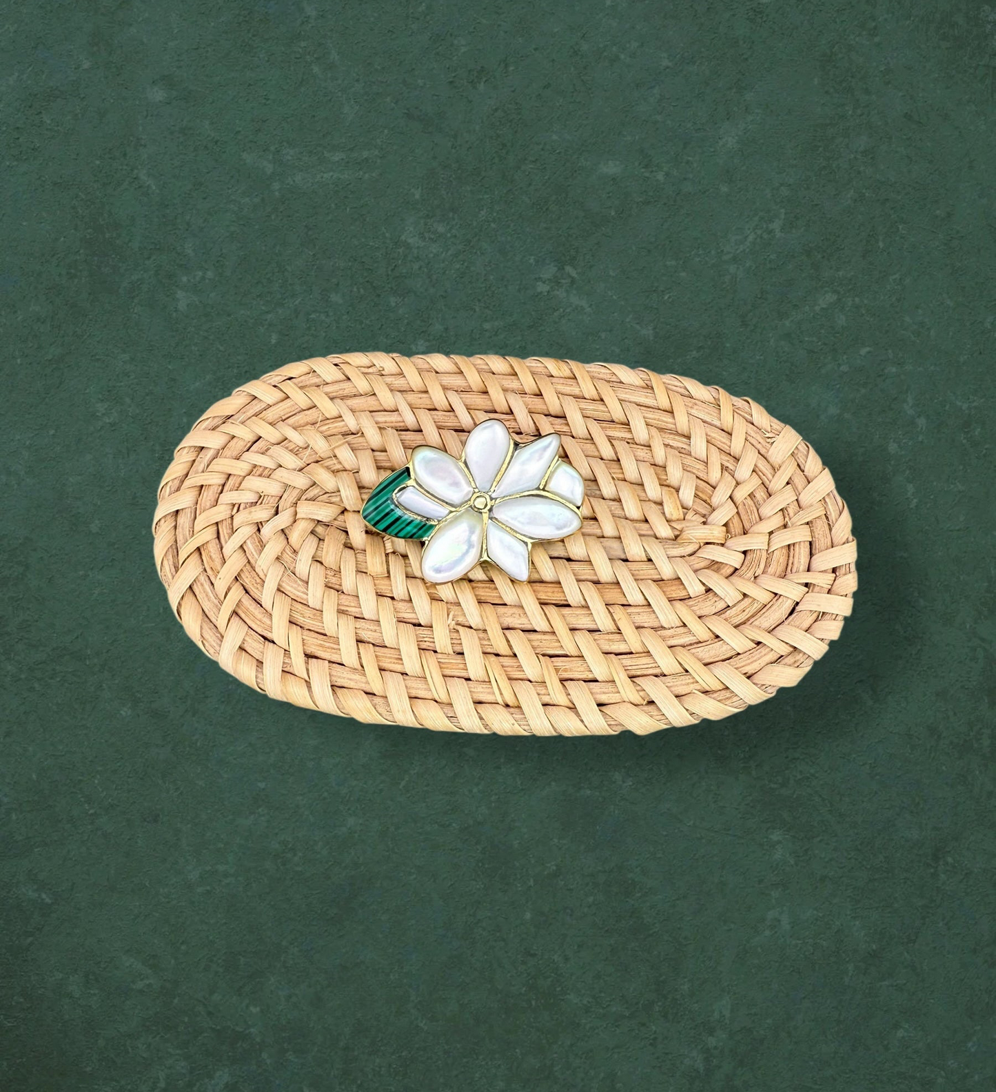 Aking Sinta Sampaguita brooch featuring hand-carved Mother of Pearl petals and a green Malachite center, displayed on a woven rattan surface against a dark green background.