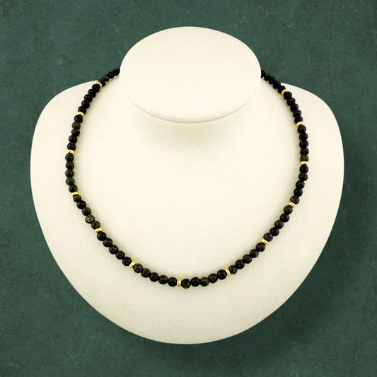 Aking Sinta Serpentine beaded necklace displayed on a white necklace holder against a dark green background; the strand features translucent lime-green gemstones accented with small, polished gold beads integrated into the pattern; no charm or extender is visible