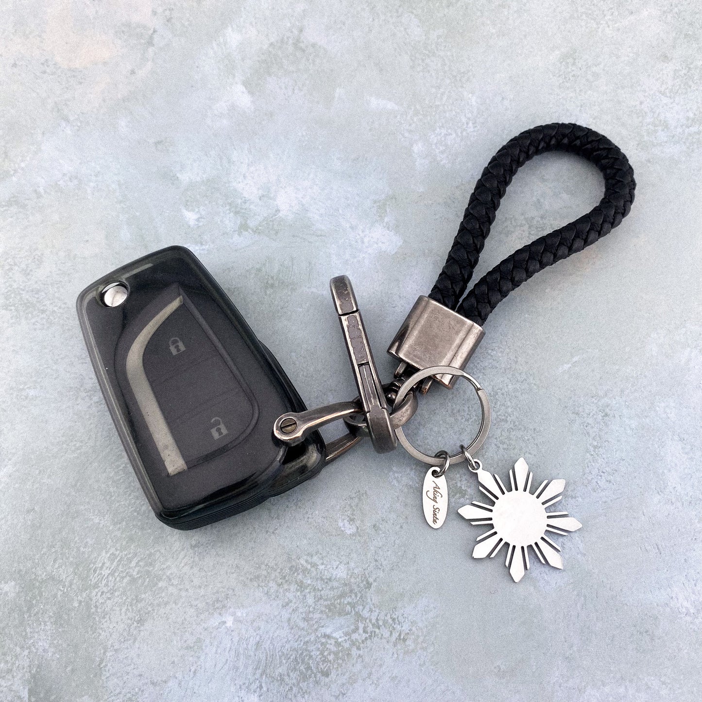 Close-up of a silver Aking Sinta Araw 8-ray sun key ring attached to a set of car keys including a black vehicle remote; a practical and stylish accessory showcasing the durability of the silver pendant