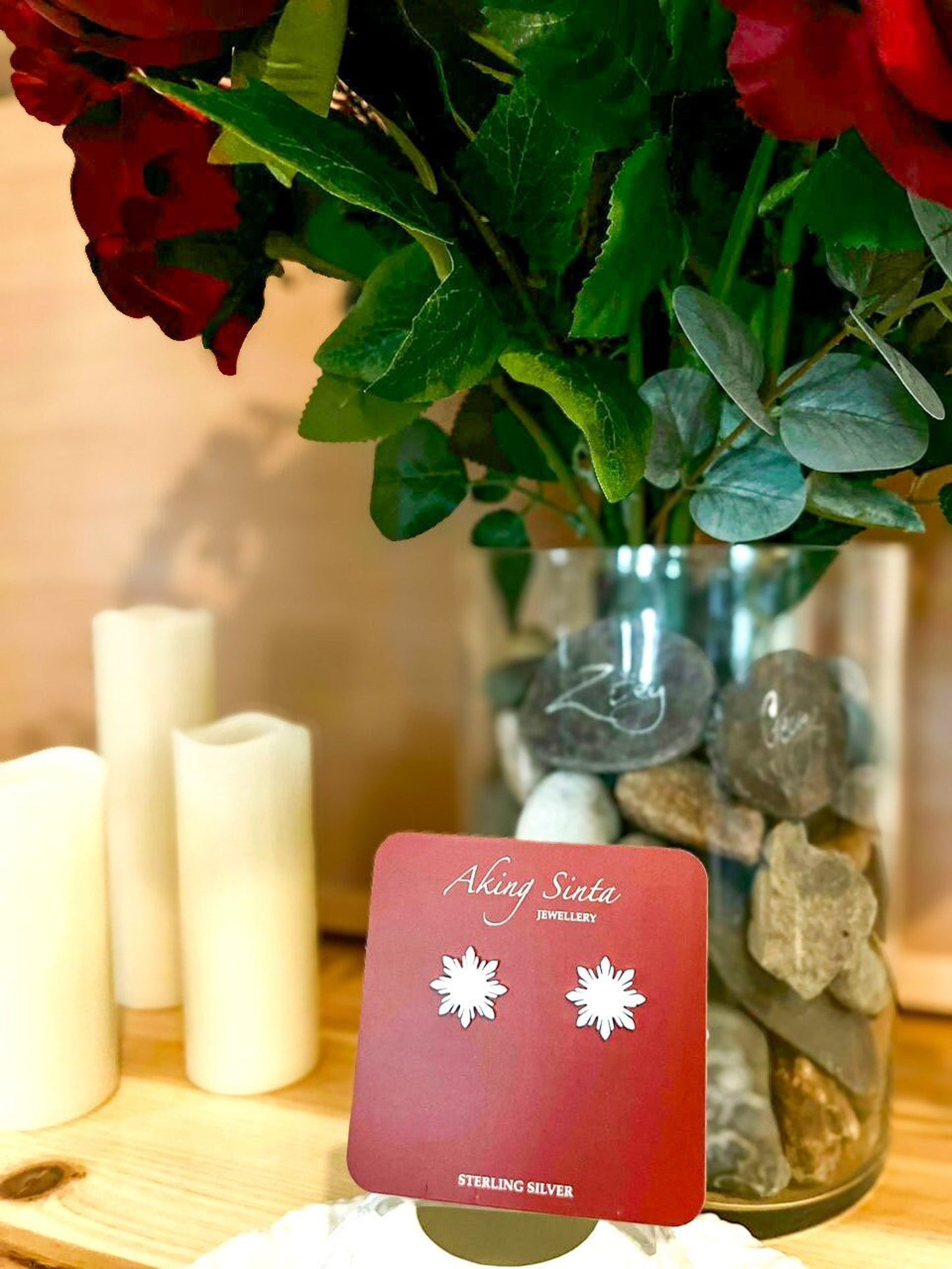 A pair of Aking Sinta 8-ray silver Araw stud earrings on a red branded jewelry card; the setting features a sophisticated lifestyle backdrop with a floral vase and a white candle for a premium gift aesthetic