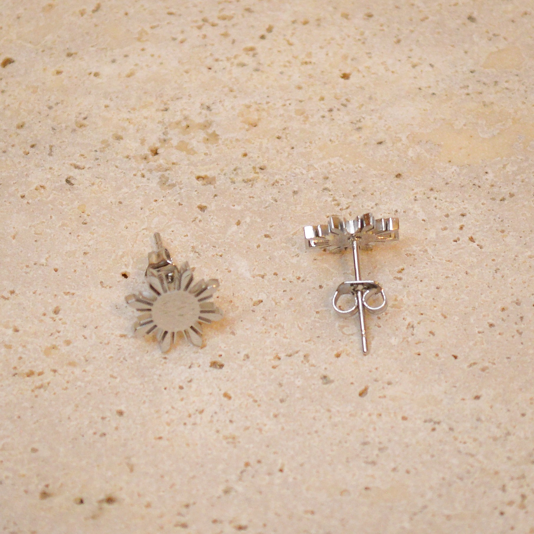 An angled close-up of a pair of Aking Sinta silver Araw stud earrings resting on a light, textured natural stone surface. One earring is shown face-up to display the eight-rayed Filipino sun design, while the other is tilted to reveal the high-polish side profile and secure butterfly back closure.