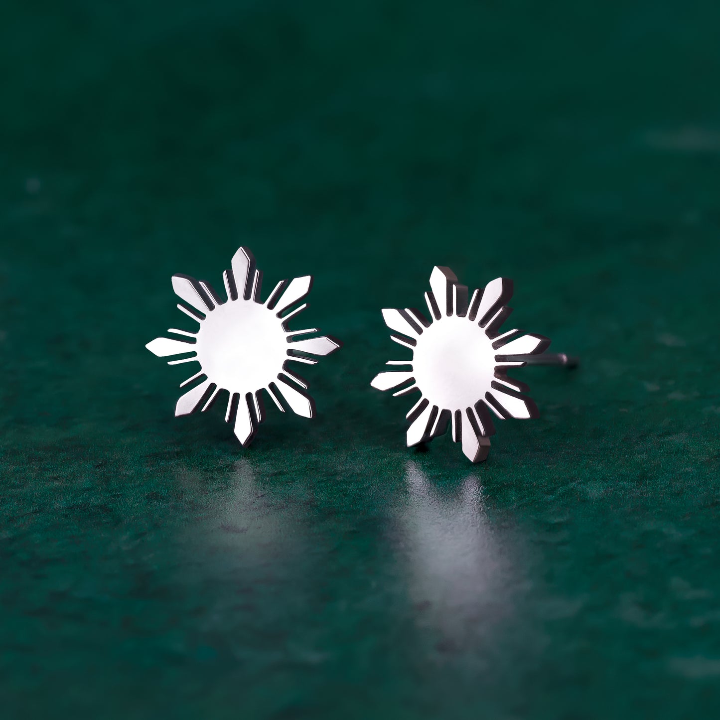 A pair of Aking Sinta silver stud earrings featuring the iconic eight-rayed "Araw" sun from the Philippine flag. The polished silver suns are displayed on a dark green textured surface, highlighting their sharp geometric lines and minimalist aesthetic