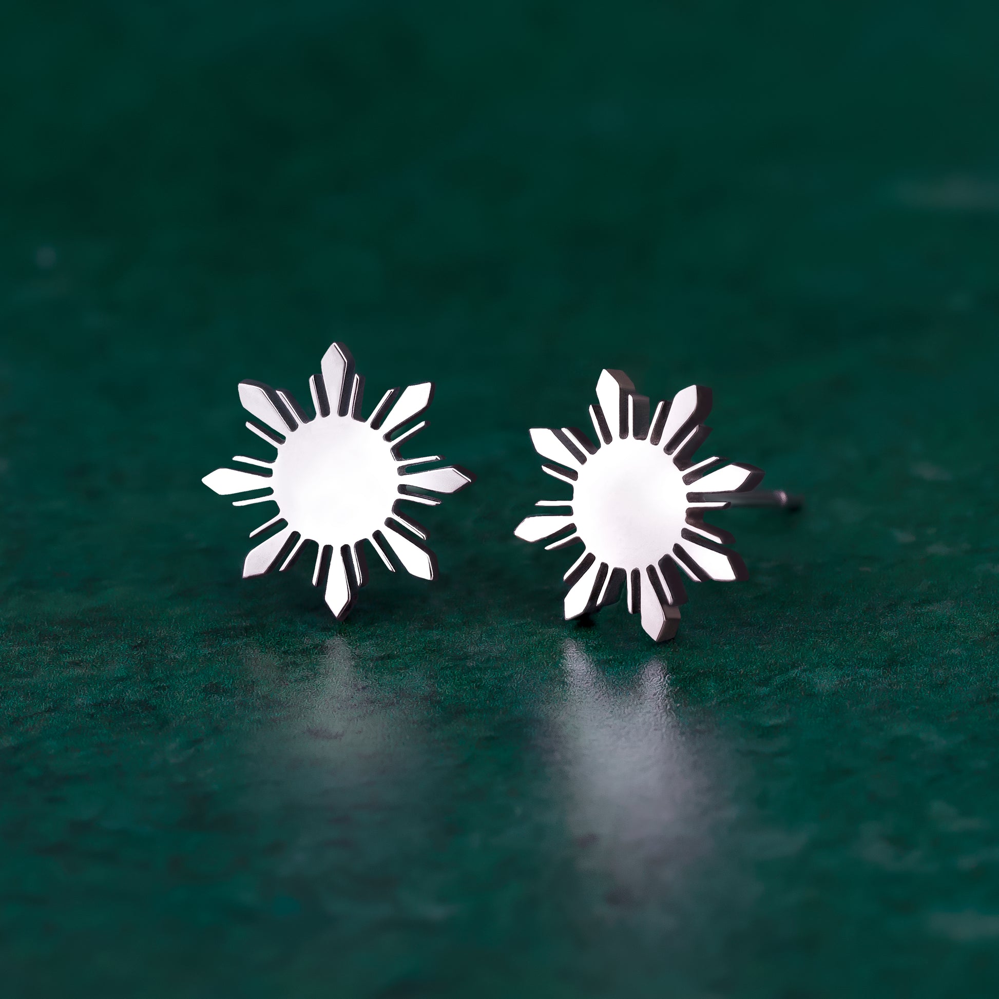 A pair of Aking Sinta silver stud earrings featuring the iconic eight-rayed "Araw" sun from the Philippine flag. The polished silver suns are displayed on a dark green textured surface, highlighting their sharp geometric lines and minimalist aesthetic