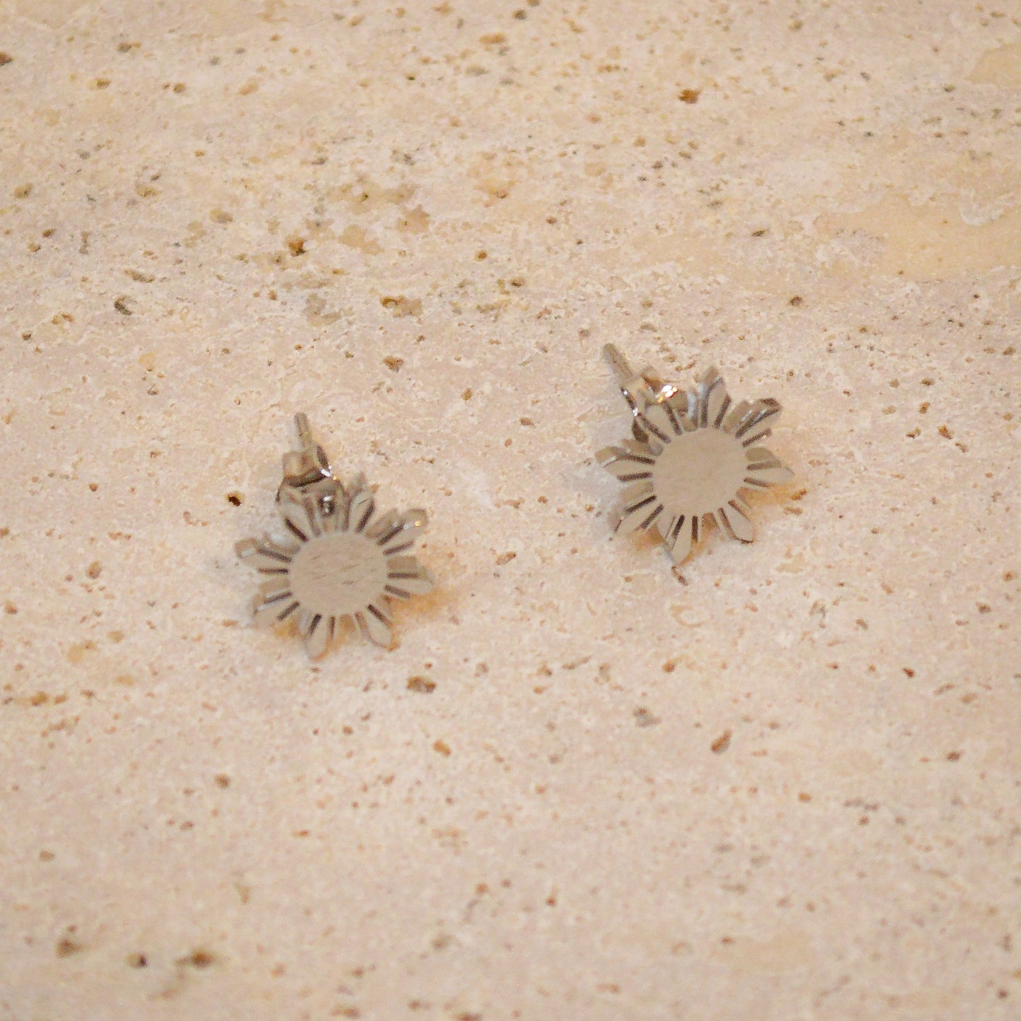 A top-down close-up of a pair of Aking Sinta silver Araw stud earrings on a light, textured natural stone surface. The polished silver finish reflects the soft light, emphasizing the sharp, geometric eight-rayed sun design inspired by the Philippine flag