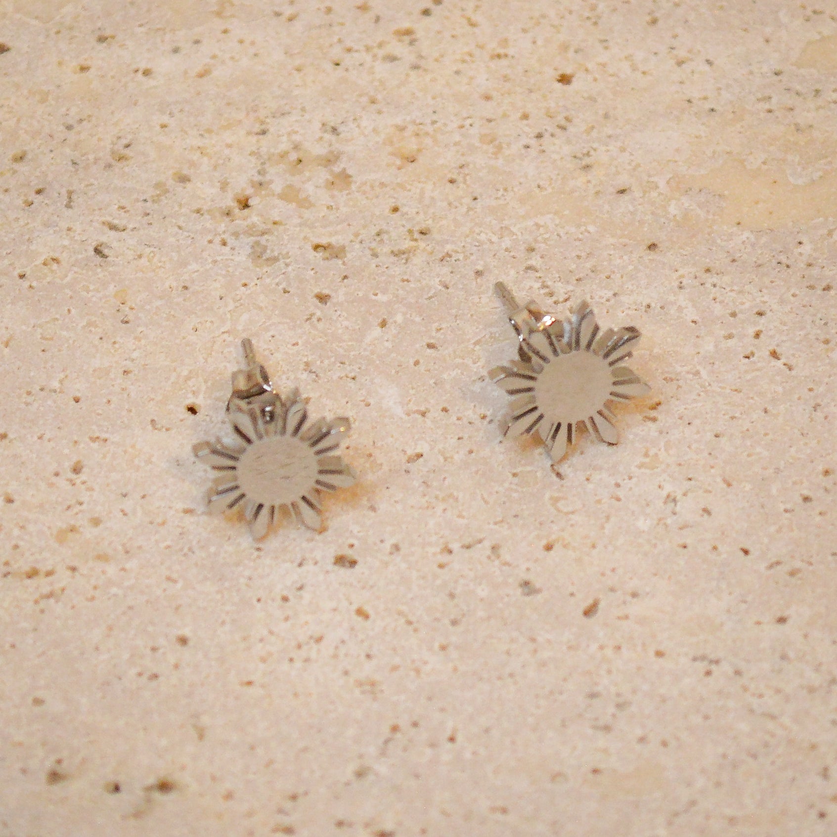 A top-down close-up of a pair of Aking Sinta silver Araw stud earrings on a light, textured natural stone surface. The polished silver finish reflects the soft light, emphasizing the sharp, geometric eight-rayed sun design inspired by the Philippine flag