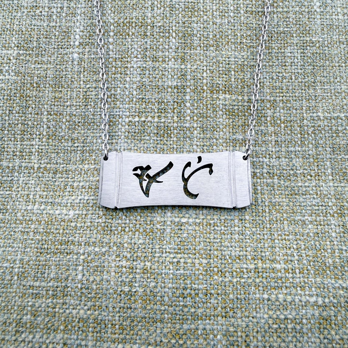 An Aking Sinta silver horizontal bar necklace centered on a textured gray-green linen background. The matte silver pendant displays a curved bamboo frame silhouette with the word "Ate" (Elder Sister) elegantly cut out in traditional script-like Baybayin characters, suspended by a fine silver link chain.