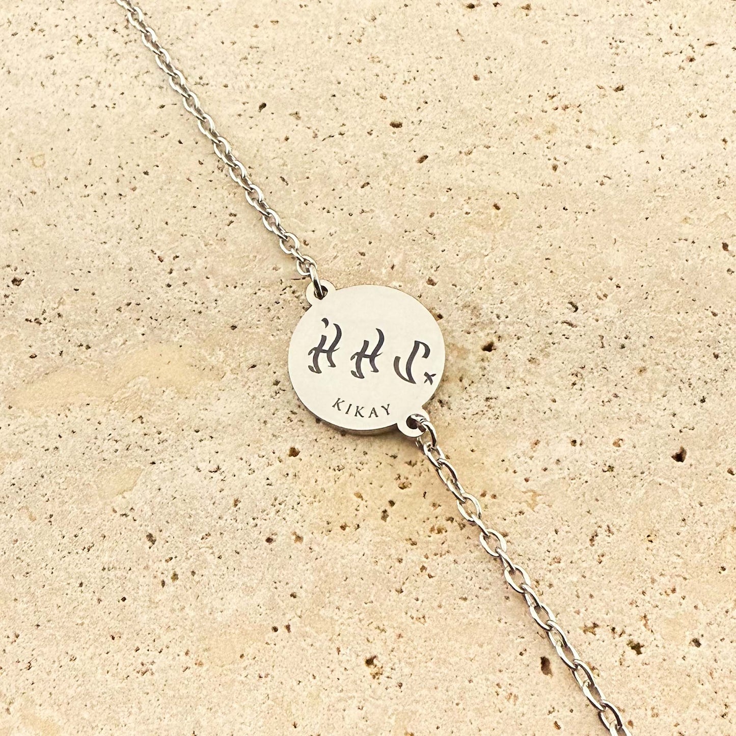 A close-up of a silver Aking Sinta coin bracelet featuring a bespoke engraving. The circular silver pendant is engraved with the word "Kikay" in traditional Filipino Baybayin script with the Latin translation below it, centered on a link chain over a light natural stone background