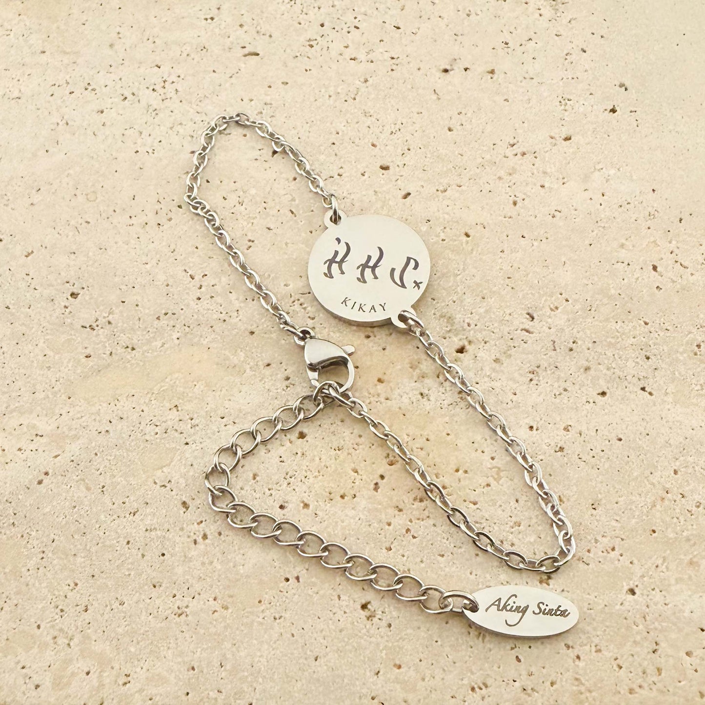 A silver Aking Sinta coin bracelet featuring a bespoke engraving. The circular pendant shows the word "Kikay" in ancient Filipino Baybayin script with the Latin text below it. The piece includes an adjustable chain with a branded "Aking Sinta" tag, displayed on a light natural stone surface
